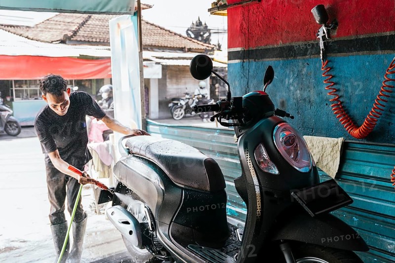 A man cleans a motorcycle using a hose at a car wash. The scene is set in an outdoor environment with sunlight highlighting the motorcycle's sleek design