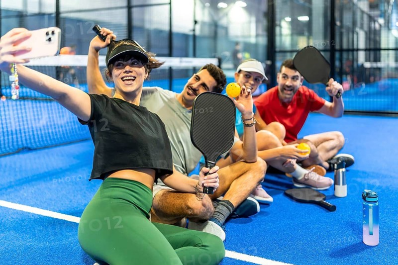 Group of smiling friends sitting on a blue pickleball court after a game, woman takes a paddle and ball selfie with smartphone, enjoying casual social fun and fitness together