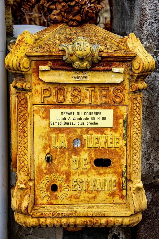 The yellow mailbox, aged and textured, stands against a rugged wall, featuring classic designs and text. France
