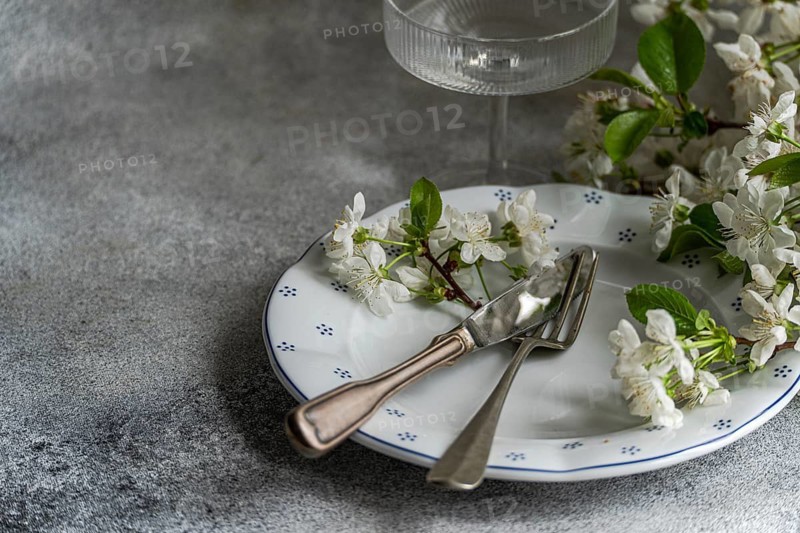 Elegant table setting featuring a white plate with delicate floral patterns, adorned with blossoming white flowers. A fork and knife rest elegantly across the plate