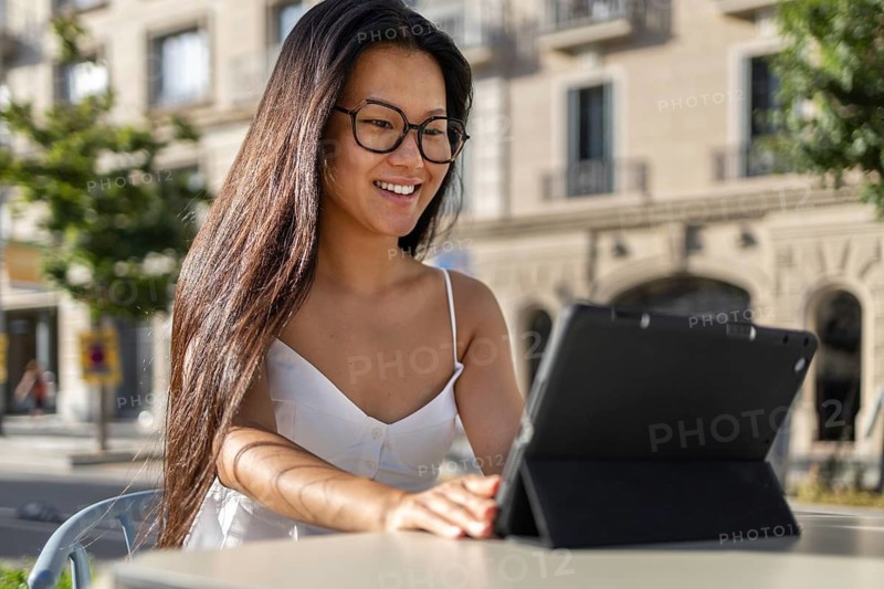 A Chinese woman with long hair and glasses enjoys a summer day in the city, sitting outdoors and using a tablet. She wears a white dress, embodying casual style