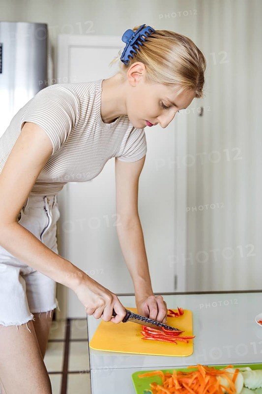 A woman is focused on chopping colorful bell peppers on a cutting board in her bright kitchen. Sunlight streams in, illuminating her meticulous work as she prepares for a meal during the day