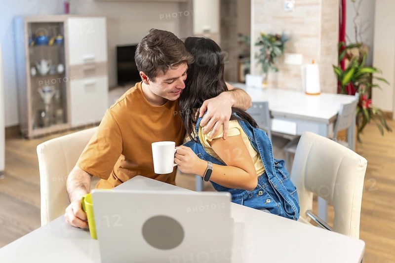 Multiethnic couple hugging at home while having breakfast and using a laptop, enjoying a cozy moment together