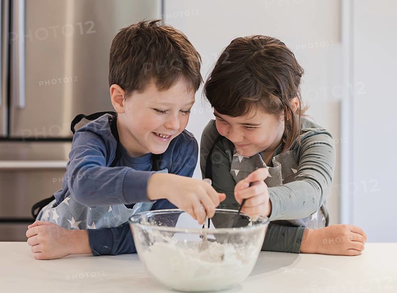 Two joyful siblings mix ingredients in a glass bowl, wearing aprons in a well lit kitchen. Their smiles capture the fun and excitement of cooking together
