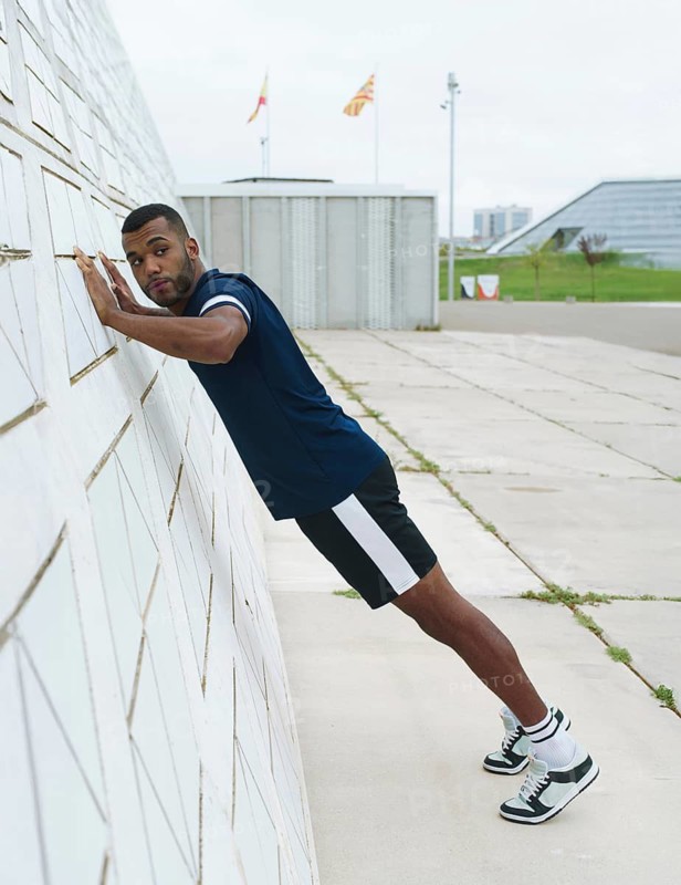 A black male athlete, wearing sportswear, performs a wall push-up outside an urban setting. The focus is on strength and fitness training