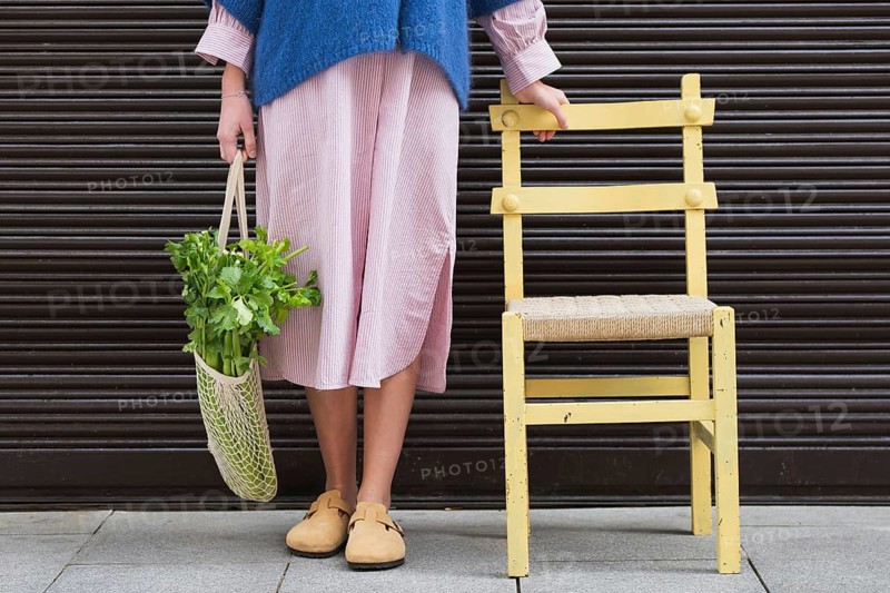 A stylish woman in a pink striped dress and blue sweater holds a mesh bag filled with fresh greens. Next to her, a vintage yellow chair on a gray sidewalk stands out