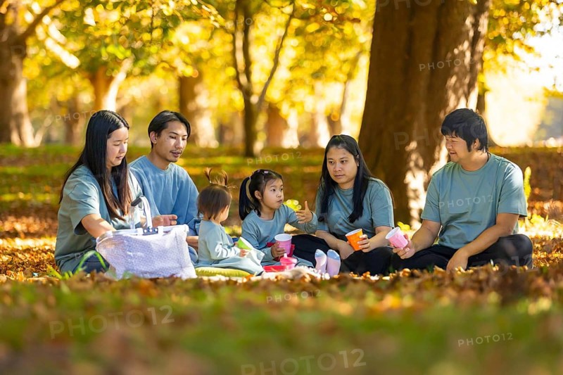 An Asian family gathers for a heartwarming picnic in a sunlit park during autumn. Surrounded by golden leaves, they share joyful moments and food, creating lasting memories
