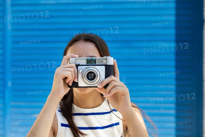 A woman embraces the urban lifestyle, capturing city moments with a vintage camera. She stands against a vibrant blue backdrop, embodying creativity and modern exploration