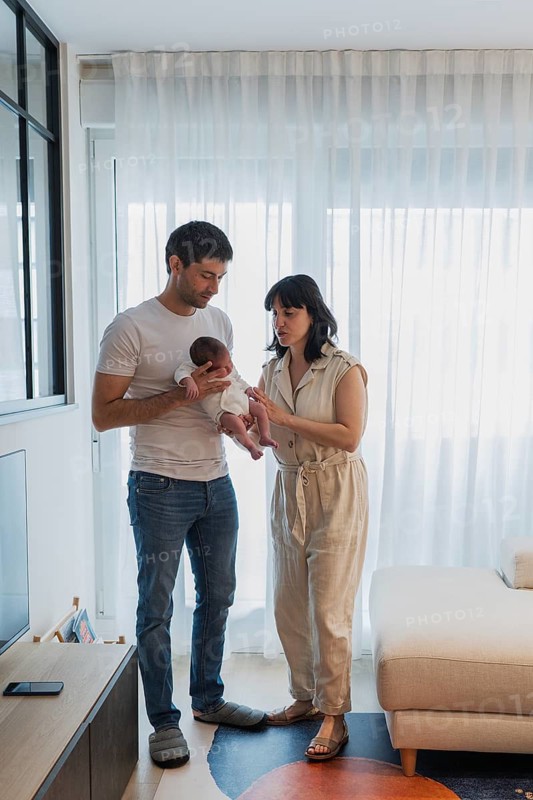 A father and mother stand together in a warm, light-filled living room, lovingly holding their newborn child. The cozy atmosphere highlights family bonds and tender moments