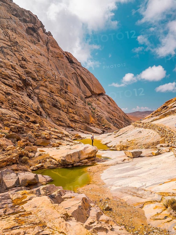 A lone adventurer stands amidst a rugged canyon, surrounded by imposing rock formations and vibrant green pools, under a bright blue sky with scattered clouds in the Canary Islands, Spain