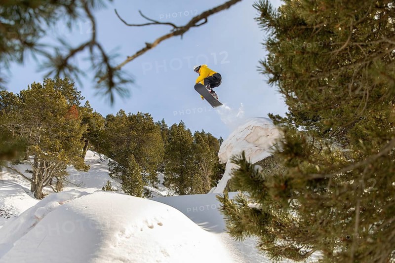 A snowboarder dressed in vibrant gear executes an impressive jump off a snowy ridge in the backcountry, surrounded by lush evergreens and clear blue skies