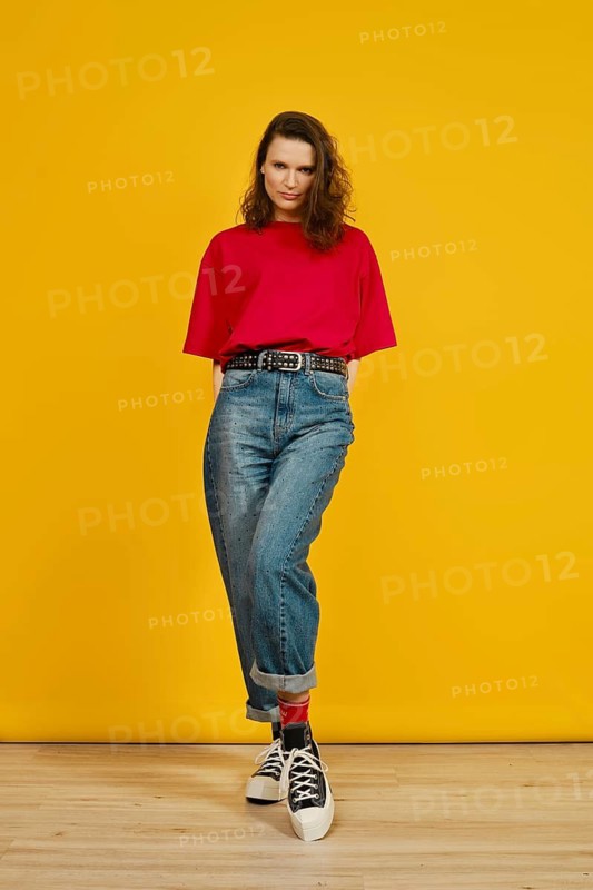 A shy woman stands with hands behind her back in a red shirt and high-waisted jeans against bright yellow background