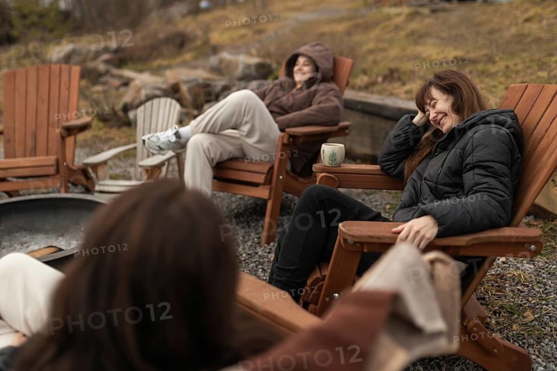 Three female friends enjoy a relaxing time together outdoors, sitting in wooden chairs and laughing around a fire pit. They are dressed warmly and the setting is a natural, wooded area