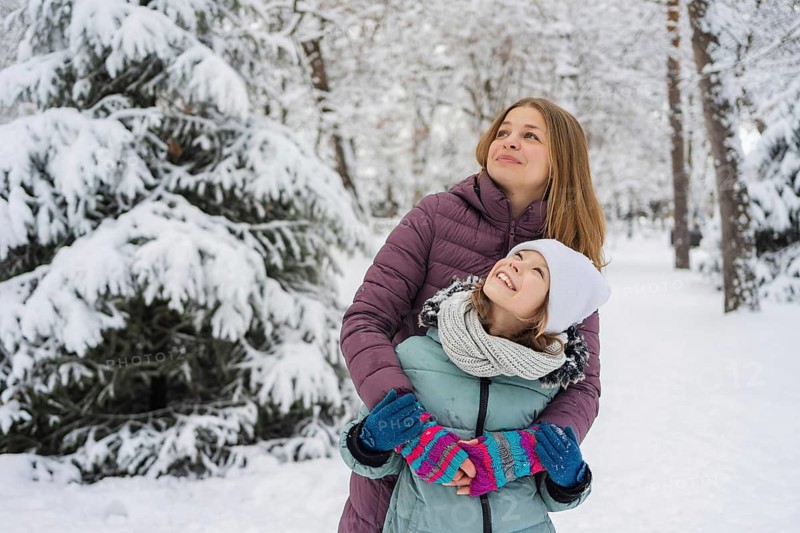 A loving mother embraces her smiling daughter in a winter wonderland. Both are dressed warmly, surrounded by snow-covered trees, capturing a joyful seasonal moment