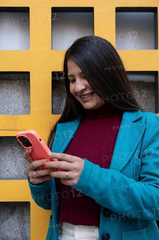 A Latin woman smiles while looking at her smartphone. She's in a blue coat and stands against a vibrant yellow grid-patterned wall, exuding a fresh and positive vibe