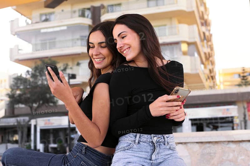 Young girlfriends sit together, joyfully interacting with their smartphones. They exude happiness and connection in a casual outdoor setting, emphasizing friendship and technology