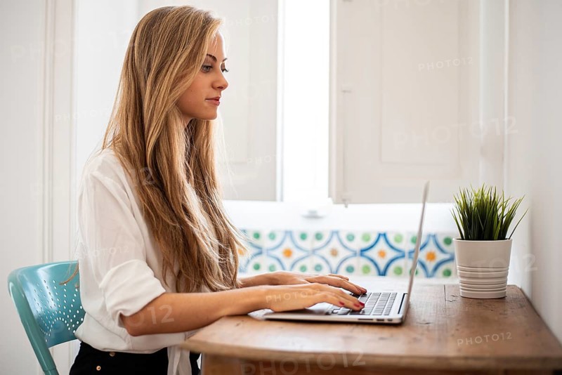A woman types on a laptop at a wooden desk in a bright room. The decor features a blue chair and a potted plant, creating a serene workspace atmosphere