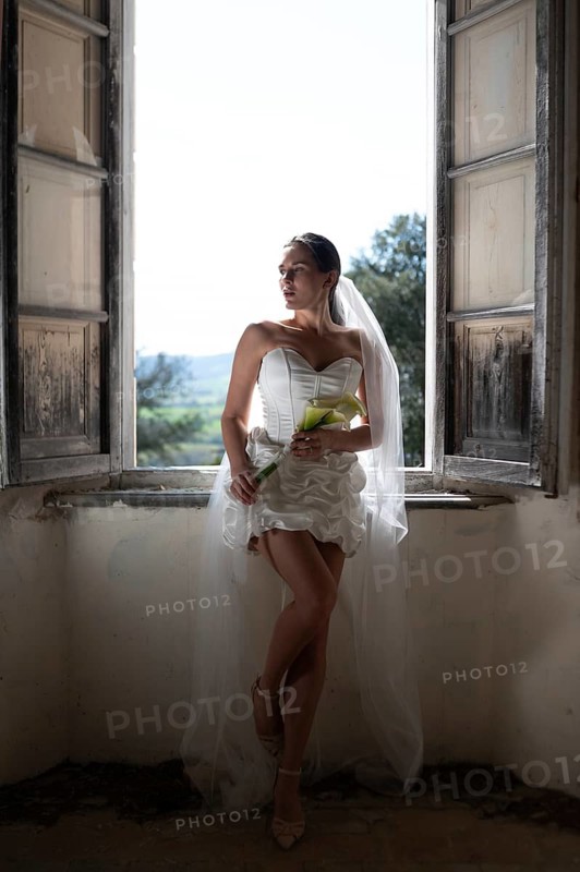 A bride in an elegant white dress and veil poses by a rustic window in an ancient villa in Treia, Macerata. The scene captures the romantic essence of an Italian wedding