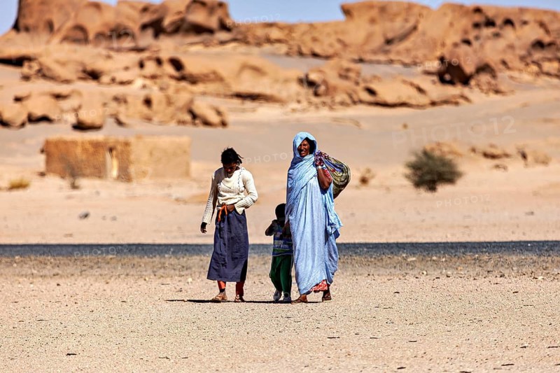 Two adults and a child in traditional dress walk through a sunny desert landscape, woman in the Sahara in Algeria