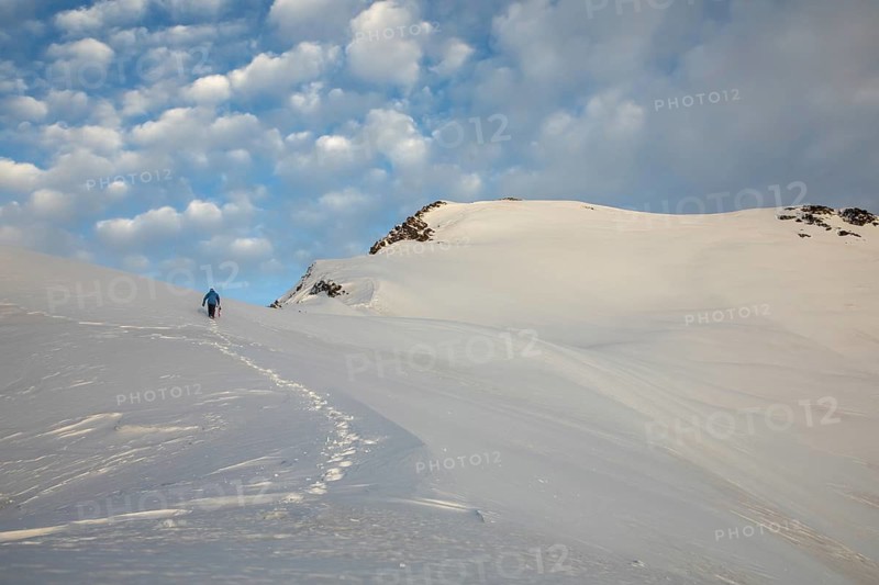 A ski tourer ascends a snow-covered mountain under a sky dotted with clouds, surrounded by untouched alpine beauty. Perfect scene of adventure and solitude