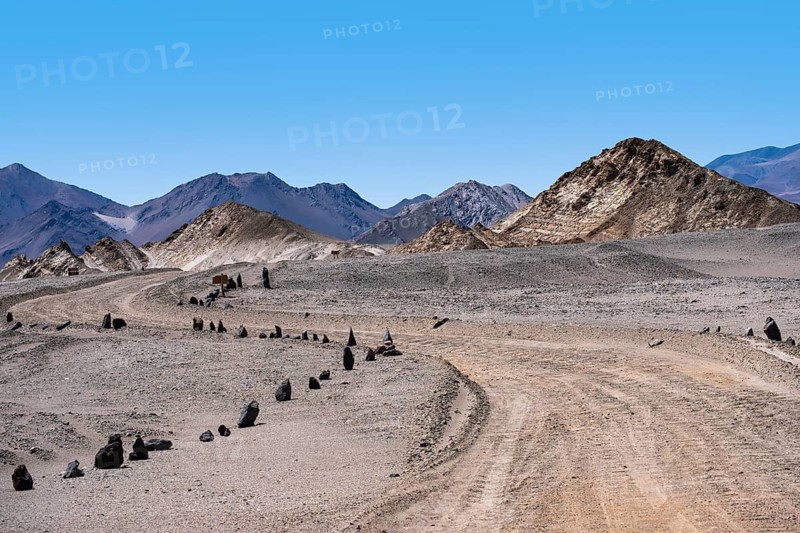 A desolate road snakes through the rocky, arid terrain of La Puna, Argentina, surrounded by rugged mountains under a clear blue sky