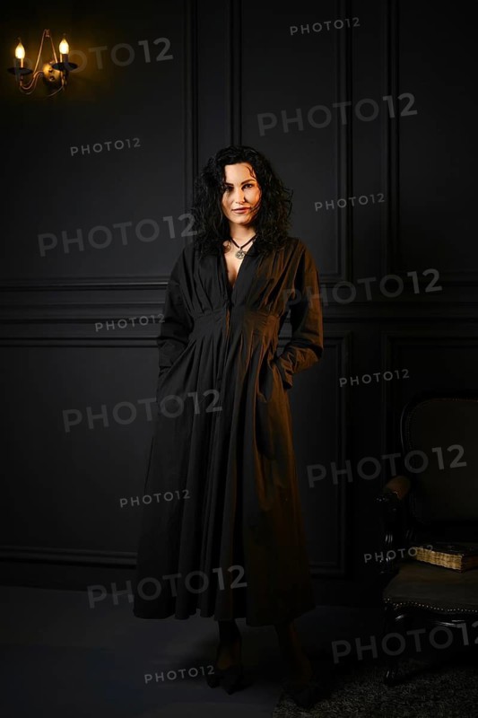 A woman stands in a dimly lit room. She wears a long black dress and has curly hair. Her hands are in her pockets as she looks directly ahead. The room features dark walls and a small light source