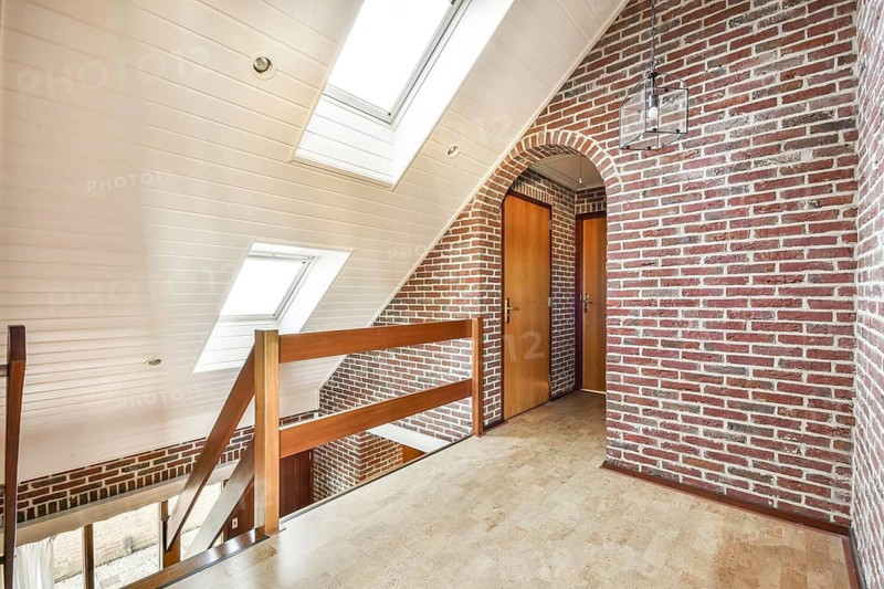 A contemporary interior design showcasing exposed brick walls, wooden railings, and natural light coming from skylights in a spacious hallway
