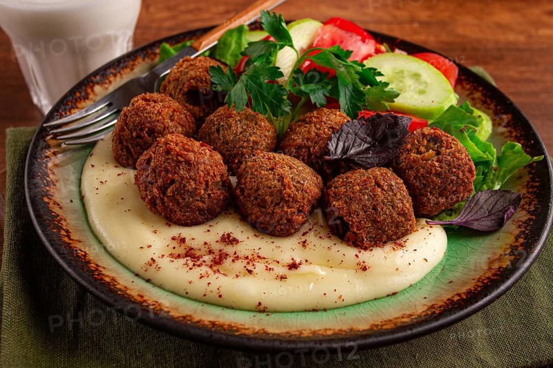 A plate holds falafel balls, tahini, salad with tomatoes and cucumbers, and a drink. The setting is warm and inviting, perfect for a midday meal with fresh ingredients