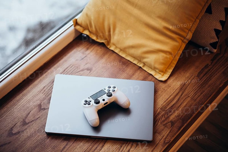 A white gaming controller rests on a closed laptop placed on a wooden window seat, accompanied by a mustard pillow, evoking a cozy and tech savvy ambiance