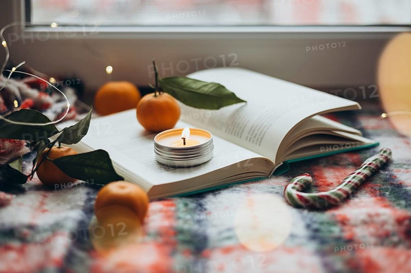 Christmas setting featuring a burning candle on an open book, surrounded by oranges and holiday lights, reflecting warmth and seasonal cheer