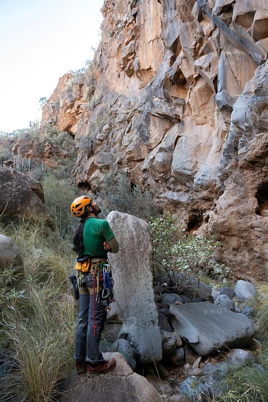 A climber stands thoughtfully at the base of a majestic rock face, surrounded by nature, preparing for a traditional climbing ascent with gear ready for the challenge ahead
