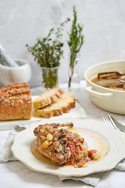 Rabbit stew is placed on a white plate next to slices of bread. There are herbs in small vases in the background. The setting is simple and shows a cozy kitchen area