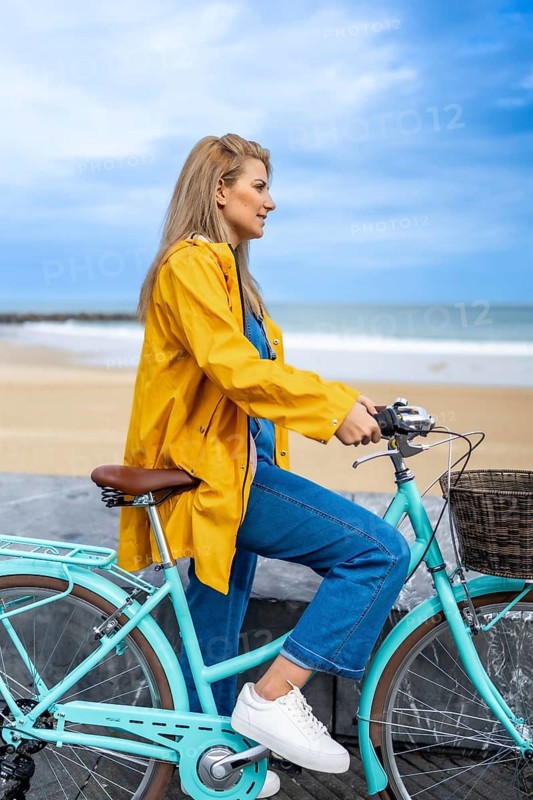 Young woman wearing yellow raincoat sitting on her light blue bicycle, enjoying the view of the sea