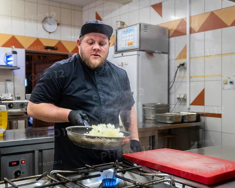 A focused chef works intently while sauteing vegetables in a busy professional kitchen. The scene reflects the chef's commitment and the lively energy of culinary creation