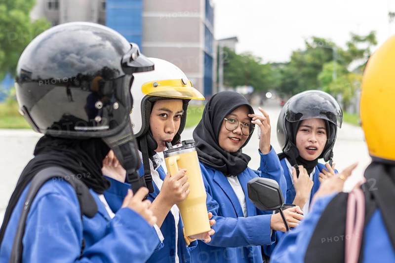 A group of Asian Muslim students wearing helmets and school uniforms gather on campus, preparing for class. They appear engaged and ready for a day of learning