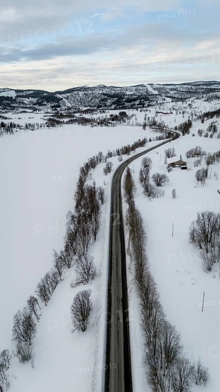 A stunning aerial view captures a road cutting through a serene snow-covered Nordic landscape. The barren trees stand starkly against the blanket of white snow, Sørreisa, Troms, Norway