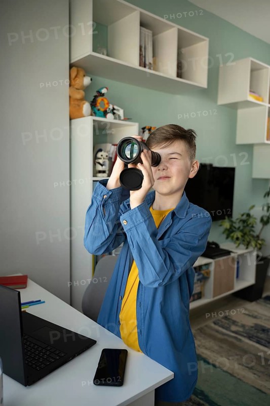Young boy enthusiastically looks through a monocular in a modern living room, displaying a sense of curiosity and exploration. Laptop and smartphone visible on desk