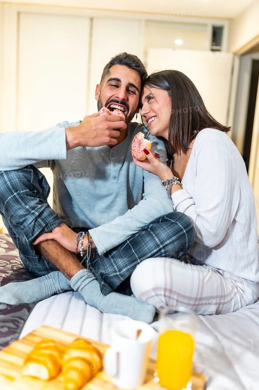 Young couple wearing pajamas, sitting in bed and eating donuts for breakfast, with orange juice and croissants on a wooden tray