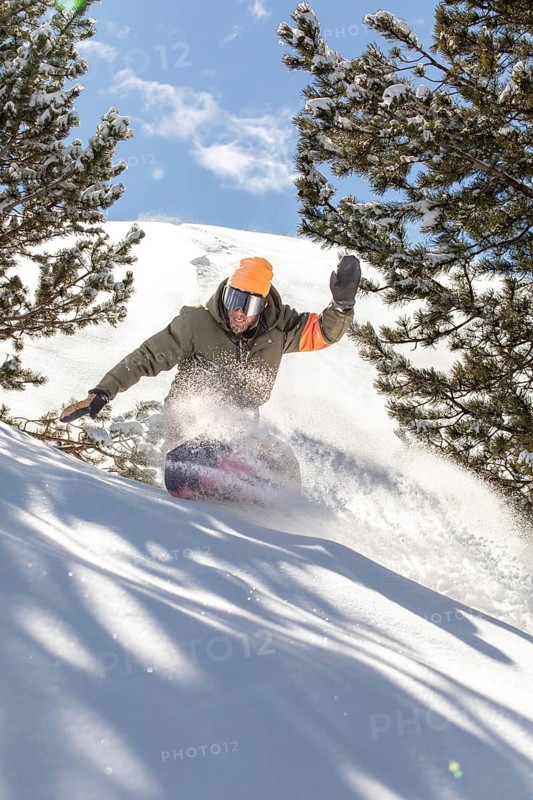 A snowboarder expertly navigates through fresh powder in the backcountry, surrounded by snow-covered trees and mountains under a clear blue sky. Perfect winter adventure