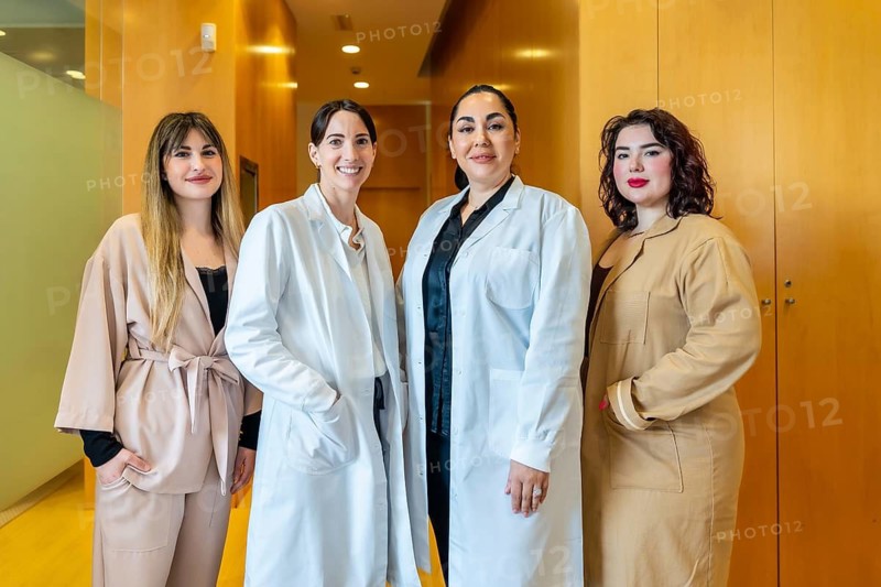 Four smiling women, two wearing white lab coats and two in professional attire, standing together in a modern clinic or beauty center hallway, representing expertise and collaborative healthcare