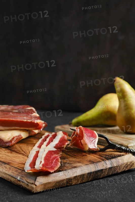 Cured duck meat slices lay neatly on a wooden cutting board alongside two ripe pears. The rustic kitchen background sets a warm atmosphere for a culinary experience