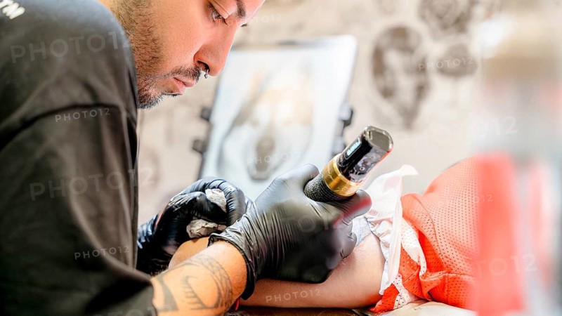 A skilled tattoo artist intently focuses on creating an intricate ink design on a client's arm, demonstrating precision and artistry in a well-lit studio setting