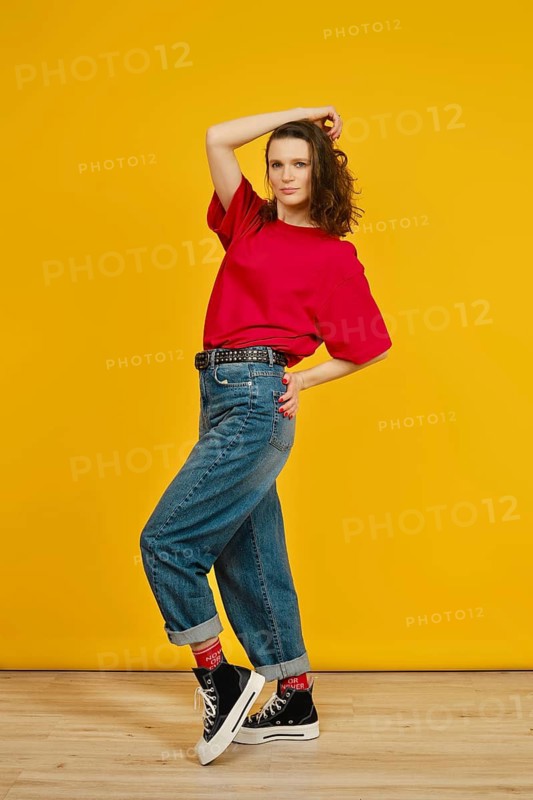 A model poses confidently in a red shirt and baggy jeans, enhancing her stylish outfit with black and white sneakers. The bright yellow wall adds a cheerful vibe to the playful atmosphere