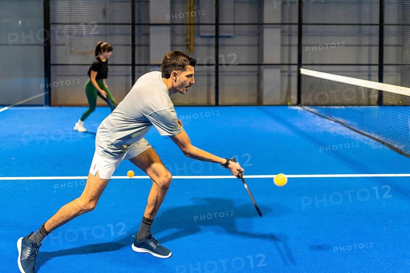 Athlete focused on hitting a yellow pickleball with a paddle on a bright blue indoor court, mid action during a competitive, energetic recreational game with intensity and movement