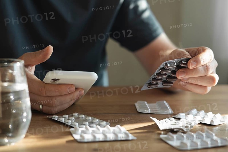 A man holds a package of pills in one hand and a smartphone in the other, sitting at a wooden table with scattered blister packs and a glass of water
