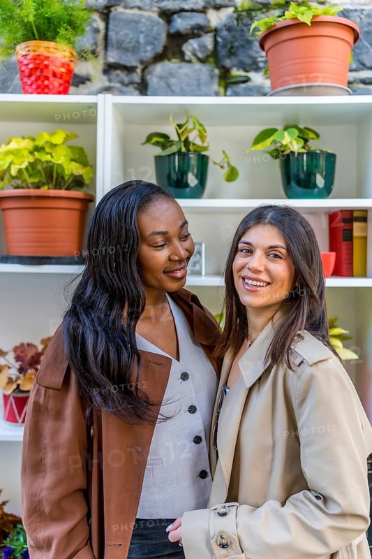 Two young businesswomen smiling together in front of a shelf adorned with vibrant plants, showcasing their friendship and professional partnership