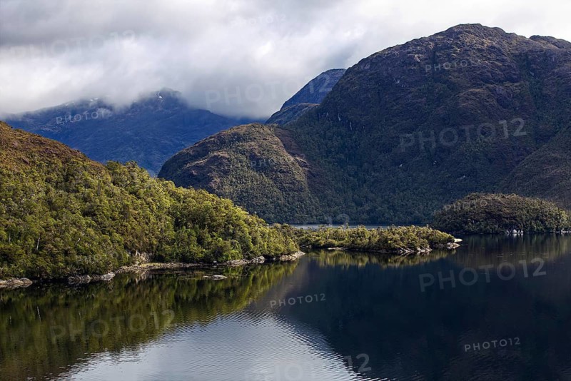 Mountain landscape with elevations of different heights, taken from the deck of a cruise ship. The left mountain is illuminated by sunlight, while the higher mountain in the right part of the picture is in shadow. Cloudy sky. The silhouettes of the mountains are clearly reflected on the calm water surface, Chile, South America