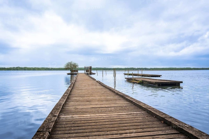 Lake azur in landes, france, offering a tranquil natural escape with a long wooden pier extending over calm blue water, reflecting a cloudy sky and distant green shoreline