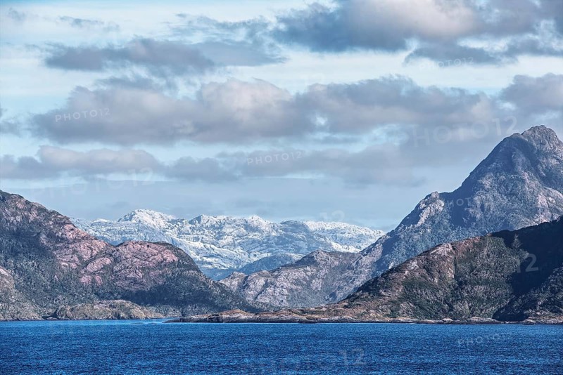 Extensive mountain landscape with mountain ranges of different heights. Behind the darker front mountains, there is a very light, gray massif that looks like snow-capped peaks from afar. Above a blue sky with passing clouds, Chile, South America