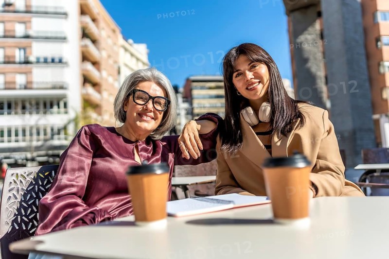 Two smiling women, an older mother and younger daughter, sitting at an outdoor cafe table with coffee cups and a notebook, enjoying time together on a sunny day in the city